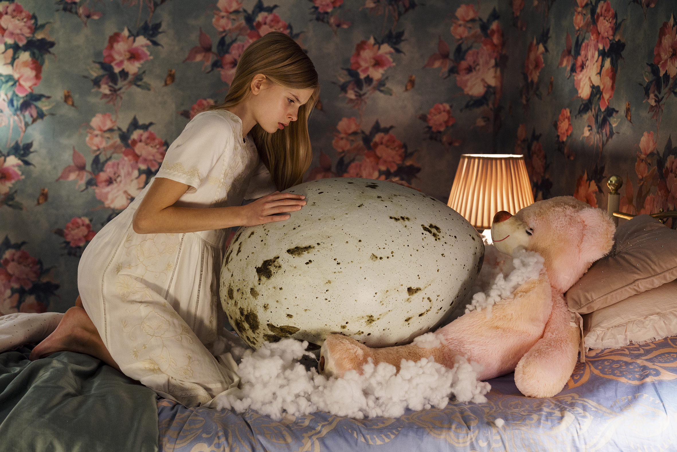 Nuori tyttö sängyllä valkoisessa yöpuvussa ison linnunmunan vieressä.